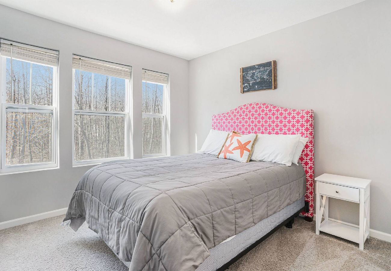 Queen bedroom with gray bedding, pink patterned headboard, white nightstand, and large windows overlooking trees