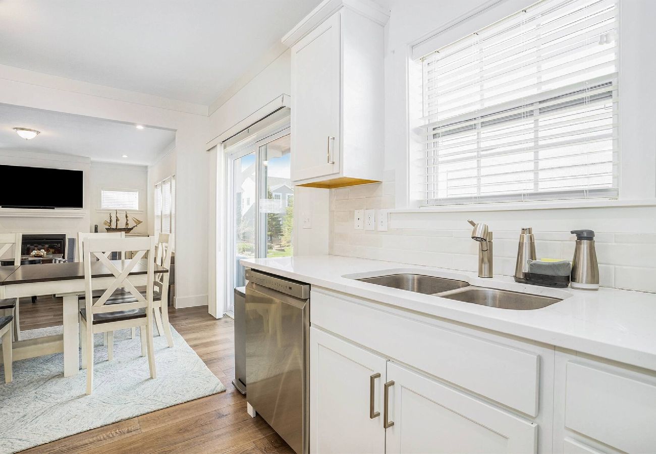 White kitchen counter with double sinks and stainless dishwasher, opening into a dining area and living room