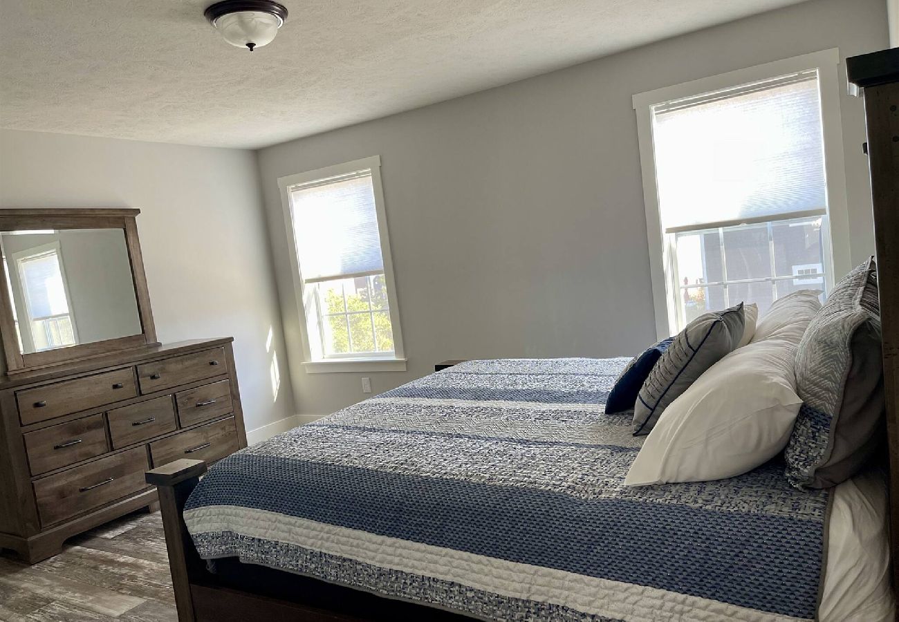 Primary bedroom with wooden dresser, king bed with blue and white bedding, and two bright windows