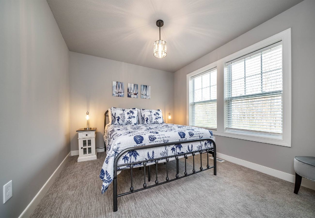 Bedroom with a black metal-frame bed,coastal-themed bedding, bedside table with lam, and large windows 